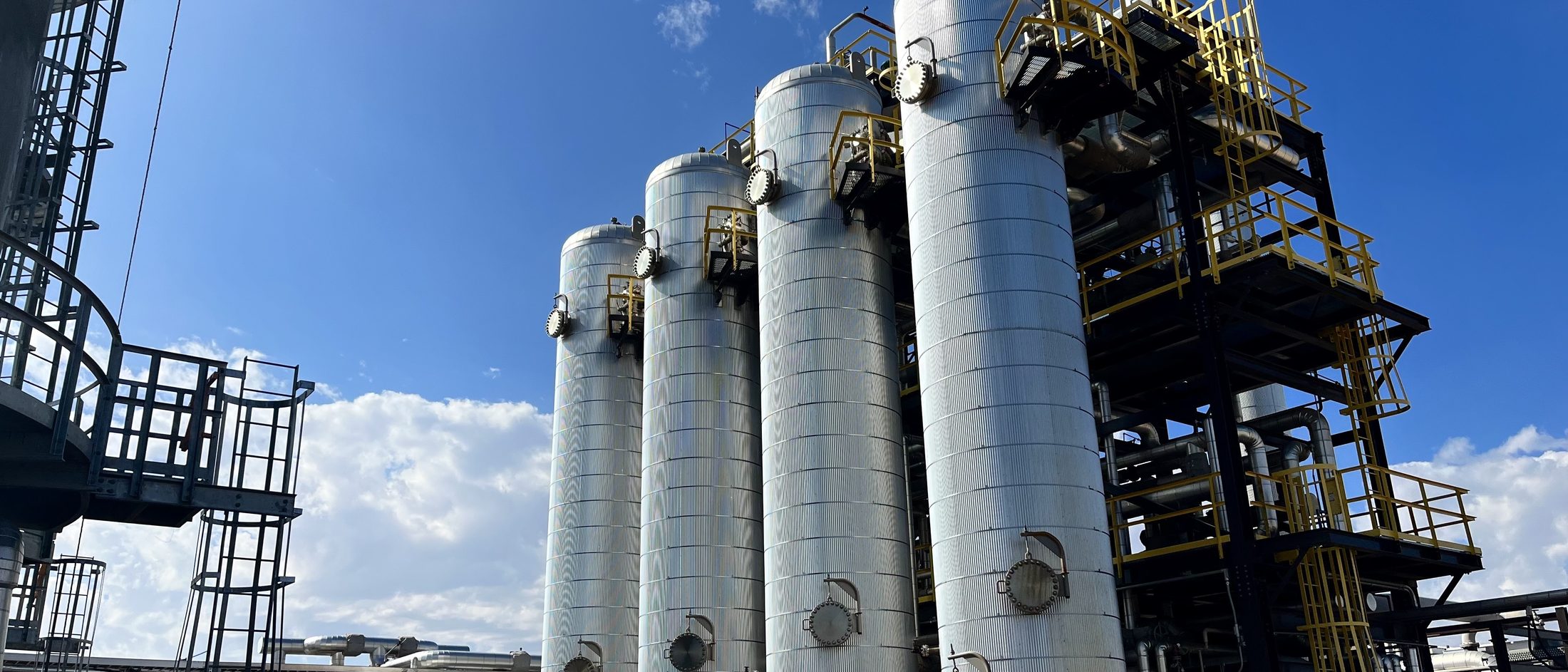 A row of four tall insulated process vessels beside a steel structure under a clear blue sky at Project Roadrunner